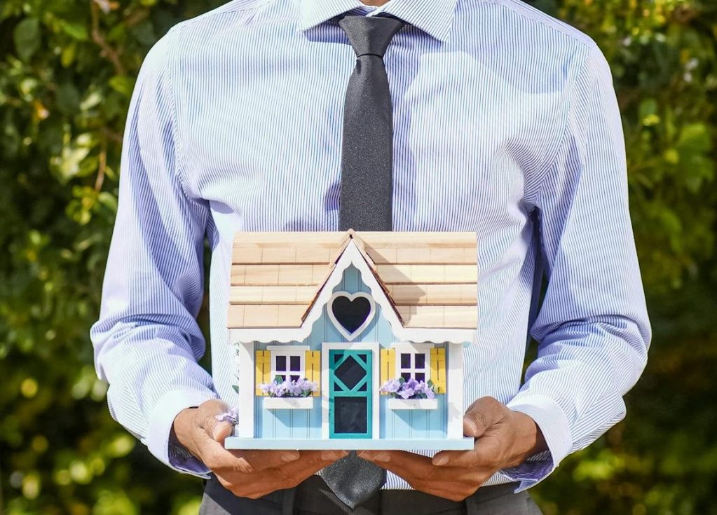 Smiling real estate agent holding a model house in an outdoor setting.